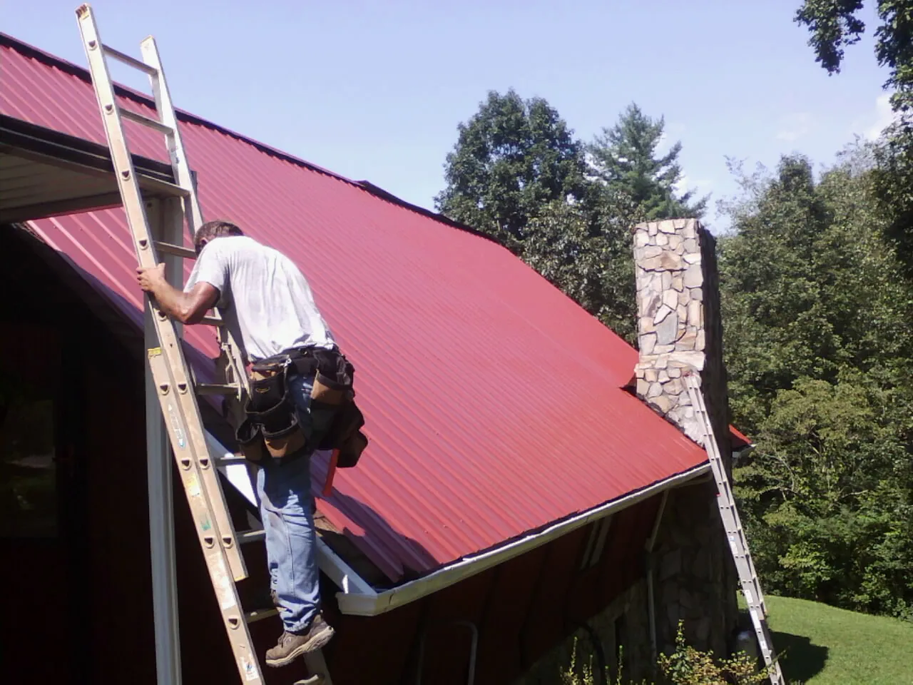 Roofer on a ladder beside a red metal roof and stone chimney — Soffit & Fascia Repair by Roofing Brothers in Austinville